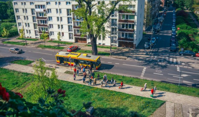 Zdjęcie przedstawia ulicę z widoku z okna. Znajduje się na niej autobus i obok na chodniku grupa ludzi.