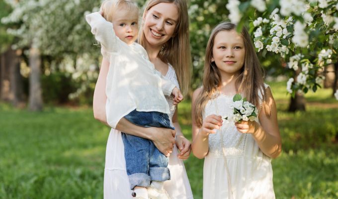 Przedstawia trzy osoby - matkę, która trzyma dziecko na rękach oraz dziewczynkę obok nich. Na tle trawy i białokwitnących drzew